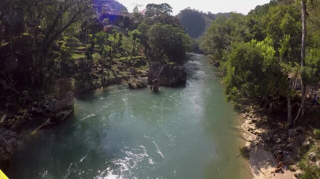 People sit on the side of a tropical river in Semuc Champey, Guatemala. Kids climb up rocks with inflatable tubes