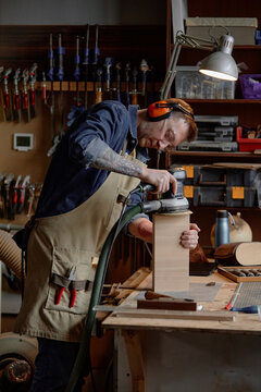 Man using electric sander on wooden block in workshop filled with tools and gadgets. Concentrating while standing at workbench in woodworking setting with shelves in background