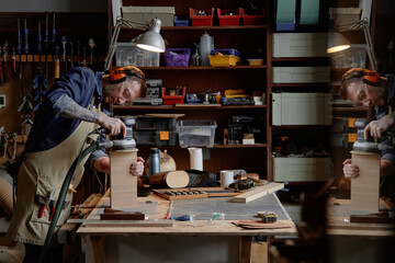 Skilled woodworker sanding a wooden piece in well-organized workshop while wearing protective gear. Surroundings include various tools and materials neatly arranged