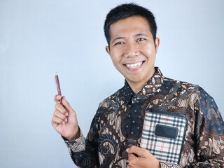 Adult Asian man wearing batik clothes smiling while holding a book and pen