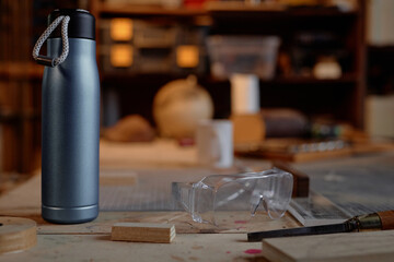 Metal thermos standing on cluttered workshop table featuring tools, safety goggles, and materials. Background showcasing various objects, adding to the cluttered workspace setting