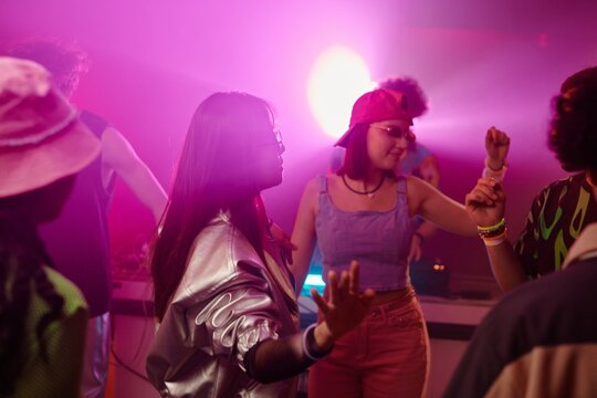 Young Asian woman with dark long hair dancing among happy intercultural friends in stylish attire in neon lit club