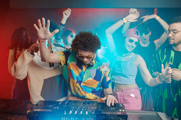 Young ecstatic male deejay with raised arm mixing sounds on soundboard while making music for...