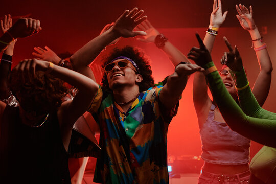 Young excited man in stylish apparel and trendy sunglasses keeping his arms raised while dancing at disco party among his friends