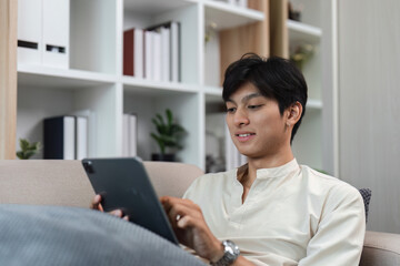 Young Man Relaxing on Sofa in Modern Living Room While Browsing Social Media on Tablet