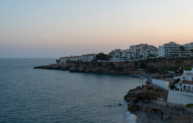 A tranquil dusk view of Nerja's coastline, featuring cliffside buildings and the calm Mediterranean Sea. Perfect for travel, sunset, and coastal themes highlighting Spain's beauty.