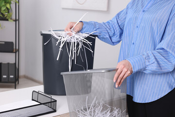 Obraz premium Woman putting shredded paper strips into trash bin at white table in office, closeup
