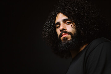 Fototapeta premium Closeup portrait of a Hispanic man with curly, shoulder-length hair and a thick, well-kept beard, black background