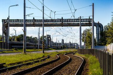 Fototapeta premium Railway tracks curve under industrial pipelines and overhead wires in an urban setting. The image highlights transportation and industrial infrastructure beneath a clear blue sky.