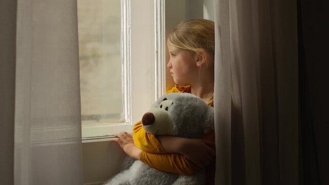 Sad little girl child kid daughter sitting window hugging holding soft teddy bear toy divorce bored abuse victim innocence depressed alone pensive solitude worry despair frustration psychology trauma