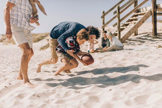 Father and son playing football on the beach with family in the background