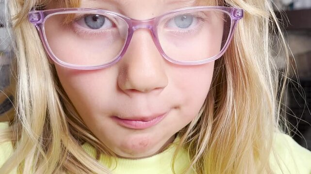 A young girl with long blonde hair holding a cherry by the stem near her mouth and eating another cherry. The background is an indoor setting with blurred grey walls