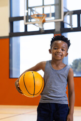 Holding basketball, smiling boy standing in school gymnasium with hoop in background