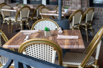 Multiple empty bistro patio dining area with four place settings on wood tables, white rattan chairs, cutlery wrapped in beige cloth napkins, and small wood greenery holders with green cactus. 