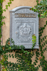 A silver metallic painted post box with a slot at the top for letters mounted on a yellow stucco exterior wall of a residential building. The mailbox has two decorative angelic cherub figures on it.