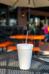 A large white nonrecyclable to-go drink cup with a plastic lid and straw on a black metal patio table. The polystyrene or Styrofoam insulated cup is filled with a cold soda pop drink. 