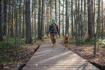 Smiling girl with cheerful smile wandering through thicket with running playful beloved pet. Joyful female backpacker together with puppy on hike, traveling, enjoying nature in joint adventure journey