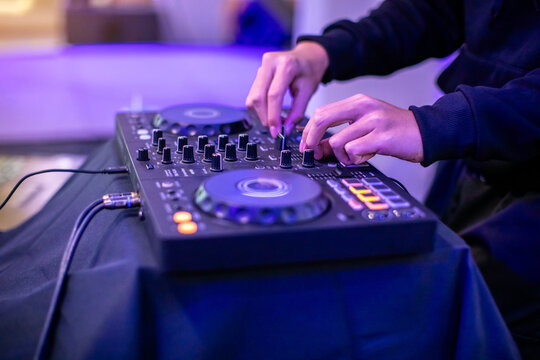 Close-up of a DJ's hands operating a modern digital DJ controller with illuminated blue and orange buttons, in a dark nightclub setting with atmospheric purple lighting.