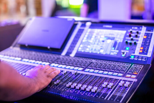Close-up of a sound engineer's hand adjusting faders on a professional digital audio mixing console during a live event with purple stage lighting.