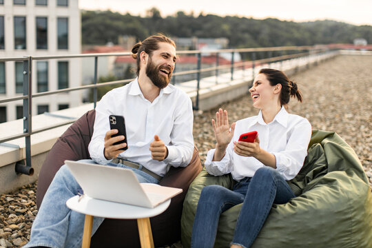 Two colleagues enjoying outdoor meeting on rooftop with smartphones and laptop. Casual work setting with laughter and bonding while discussing projects.