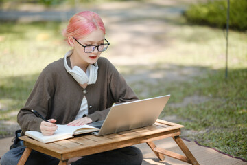 Young woman with a laptop and notes studying in the park for education in nature to relax and learn online.