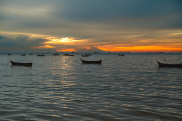 The calm sea at sunset with small boats scattered on the calm waters. The sky is a mix of warm orange and light blue, creating a peaceful and beautiful scene.
