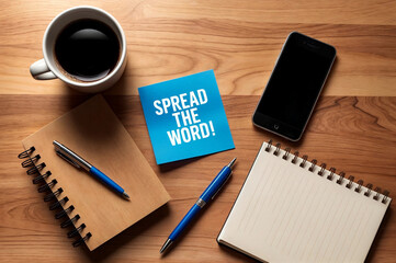 A wooden table with mobile smartphone, coffee cup and a notebook with word text phrase "SPREAD THE WORD" written on it