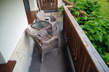 Two wicker chairs and a glass table on a small balcony with wooden railings and green plants