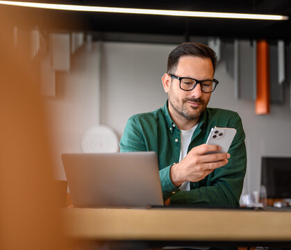 Handsome young businessman messaging online over mobile phone and working over laptop on desk in office
