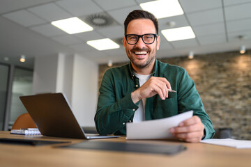 Portrait of handsome entrepreneur with laptop and documents smiling and working at desk in office