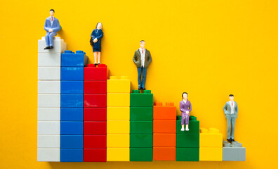 Businessmen figurines standing and sitting on top of colorful plastic building blocks forming a bar...
