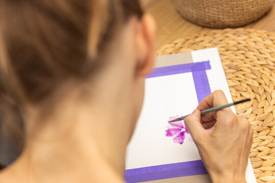 Fototapeta Artystka malująca akwarelami kompozycję kwiatową w barwach różu i fioletu. Praca umieszczona jest na podkładce z fioletową ramką. W tle widoczna część palety z kolorami oraz brązowa, pleciona faktura.