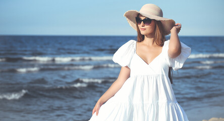 Happy smiling blonde woman is posing on the ocean beach with sunglasses and a hat