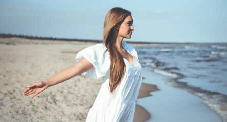 Happy smiling beautiful woman is on the ocean beach in a white summer dress, open arms