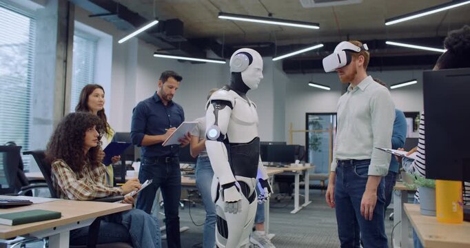 Multi-ethnic teach of engineers and programmers surrounding their new creation. Male with VR headset moving and operating humanoid robot. Checking synergy. Testing. People happy about success.