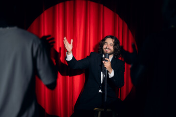 Smiling man speaking to microphone performing in comedy show open mike stand up on stage with red...