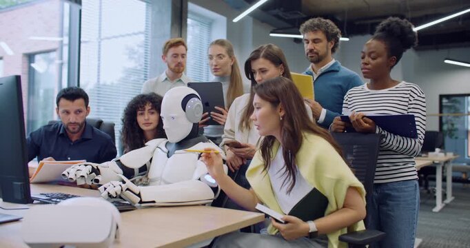 Multi-ethnic group of people surrounding robot. Humanoid working at computer. Colleagues noting everything on tablets and notepads. Woman giving commands. Testing hardware and operating system.