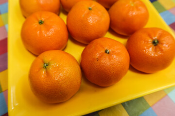 close up  top shot of nine orange citrus tangerine fruit on a yellow plate on a table with colourful table cloth