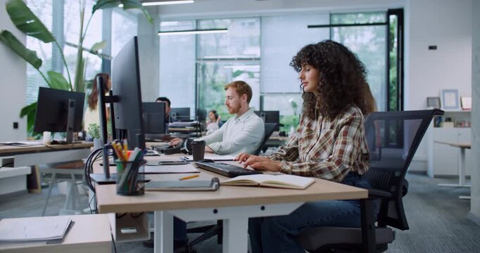 Camera moving closer to Caucasian woman while holding focus. Female with curly hair wearing checkered shirt. Looking with focus at desktop monitor. Thinking about best reply or important decision.
