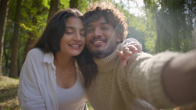 Happy multiracial couple smiling taking photo selfie filming video Hispanic woman female girlfriend make peace sign Indian Arabian man male boyfriend hold mobile phone cuddle laughing in park romance