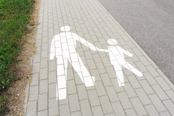 Sidewalk path background. White painted pedestrian symbol with an adult guiding a child. The cobblestone path and grass lawn with an asphalt street. Walkway view. Scenic urban pathway.