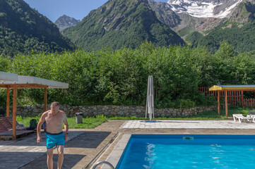 A man near an outdoor pool in the mountains with snow-capped peaks. Vacation in the mountains in summer. A man is sunbathing by the pool in the highlands. Sun loungers by the pool with blue water.