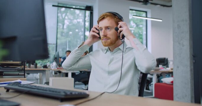 Camera focus on handsome Caucasian male sitting at desk. Putting on wired headphones with microphone. Beginning conversation with client or company. Technical support department. Smiling with joy.
