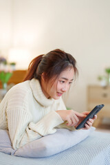 Young asian women in sweater lying on comfortable couch to enjoying with surfing entertainment on smartphone while relaxation and doing activity to spending time with slow life lifestyle at home