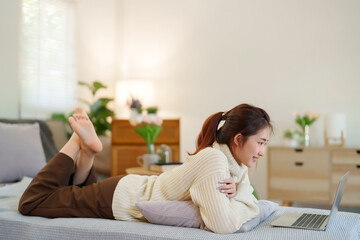 Young asian women in sweater lying on the couch to enjoying with watching social media entertainment on laptop while relaxation and doing activity to spending time with slow life lifestyle at home