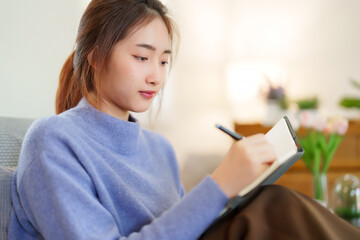 Young asian women in sweater sitting and leaning on couch to reading book and taking note for learning education while relaxation and doing activity for spending time with slow life lifestyle at home