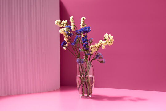 Ramo de flores secas azules y amarillas en un jarr&oacute;n con agua sobre un fondo rosa