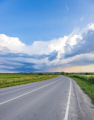 A winding asphalt road stretches out through verdant fields, leading towards a horizon framed by a blue sky dotted with fluffy white clouds.