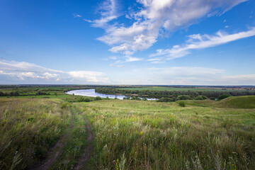 Lush green hills stretch towards a winding river, reflecting the clear blue sky, as soft clouds drift by, capturing the tranquility of nature's beauty.