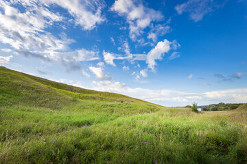 Soft green hills stretch beneath a vast blue sky, dotted with fluffy clouds, creating a peaceful...